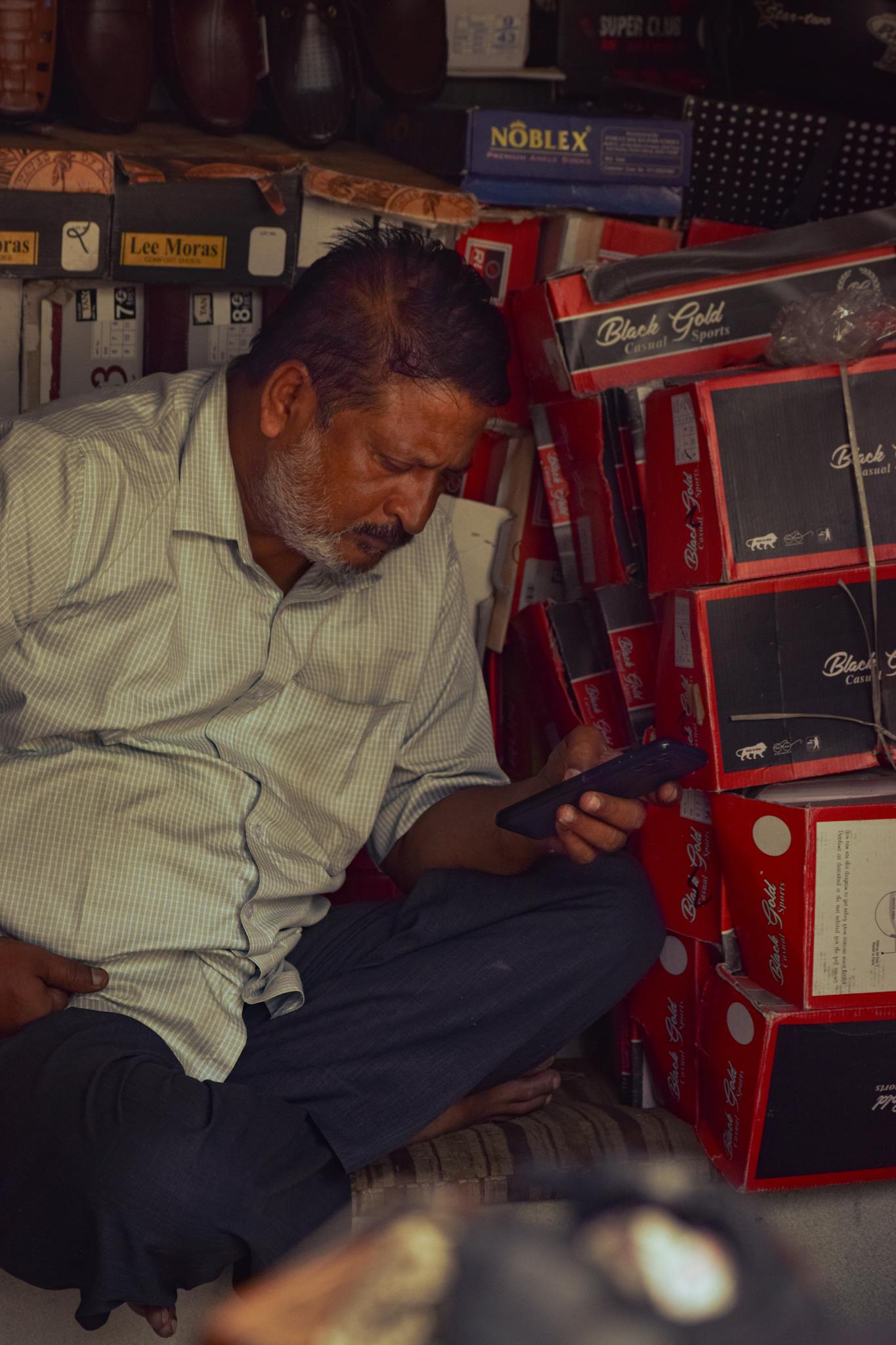A man in a small shoe shop uses a smartphone, surrounded by stacked boxes.